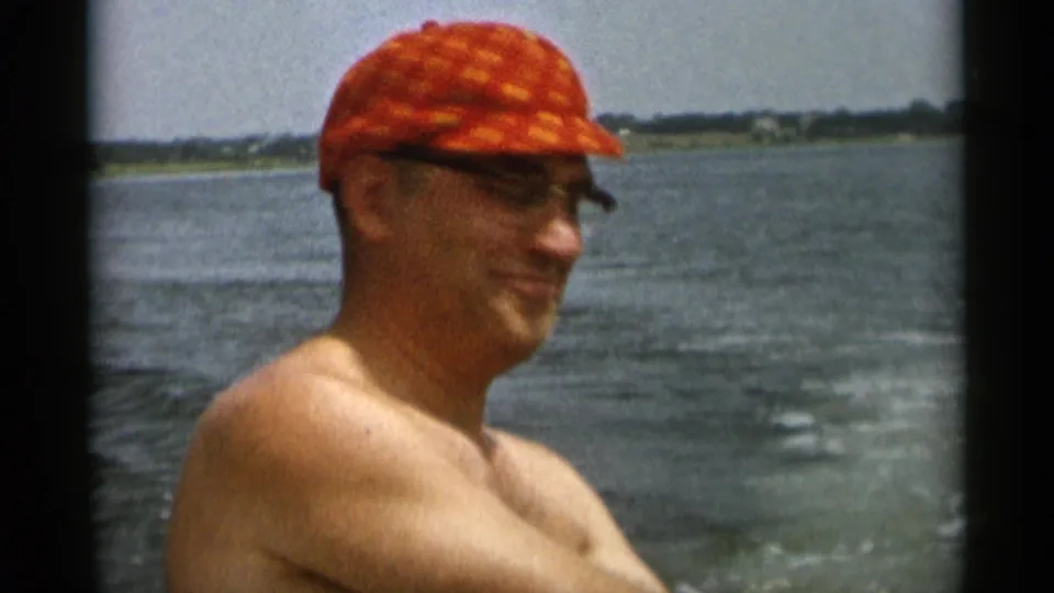 1956 Cape Cod 8mm frame showing a shirtless adult man in profile wearing glasses and a red cap beside open water.