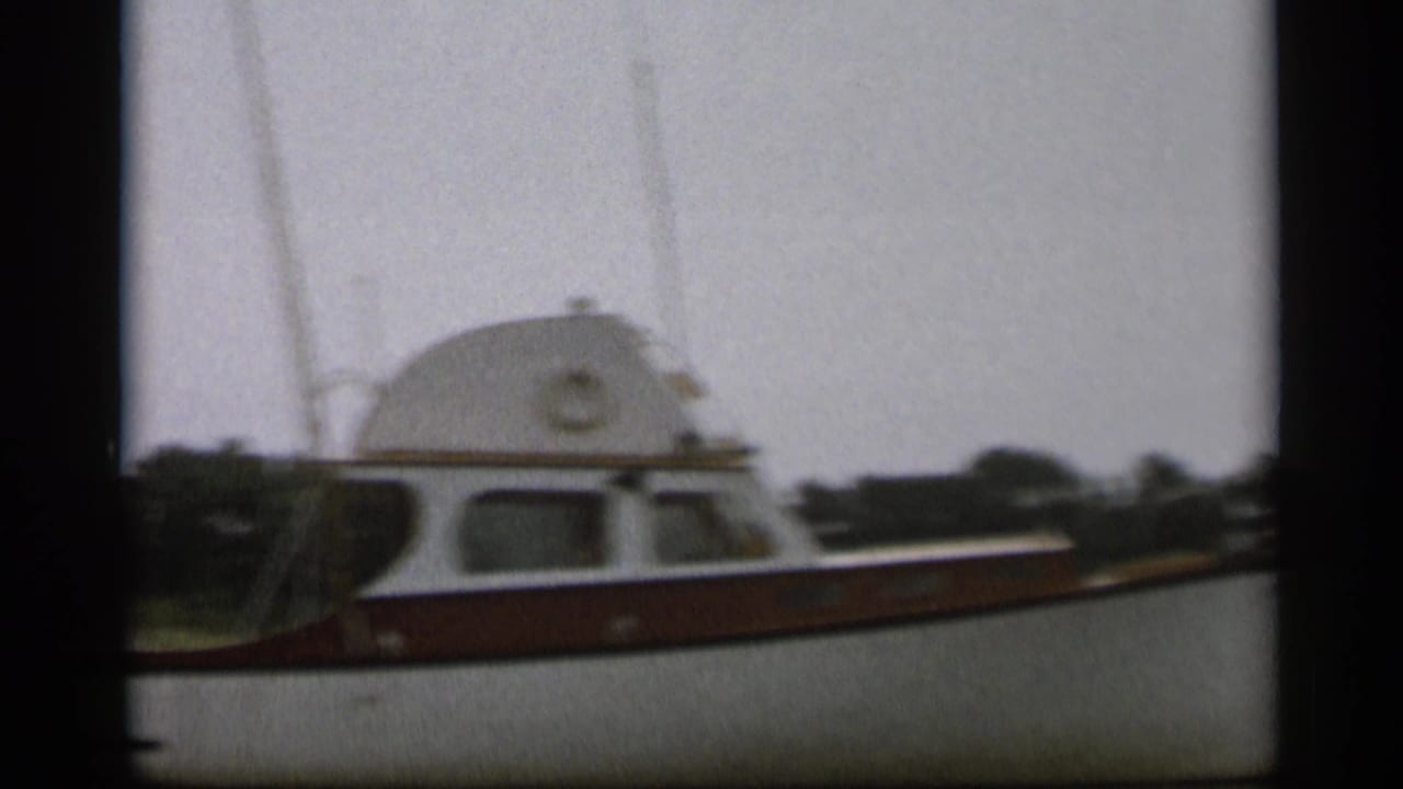 The white and brown cabin motorboat remains near the camera, with dark film borders and shoreline behind it.