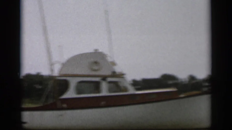 The white and brown cabin motorboat remains near the camera, with dark film borders and shoreline behind it.