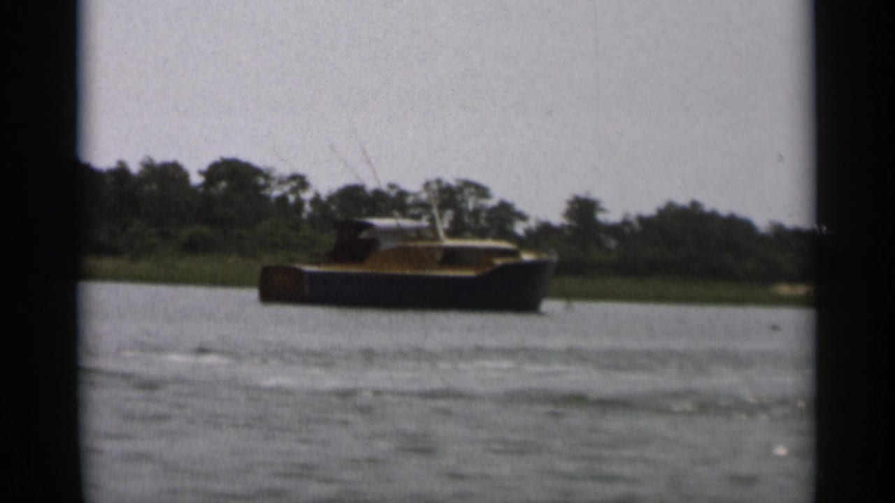 1956 Cape Cod 8mm frame showing a small dark-hulled cabin boat crossing calm water near a low wooded shore.