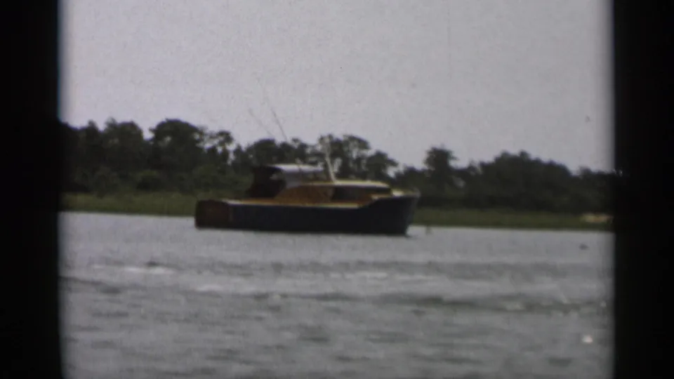 1956 Cape Cod 8mm frame showing a small dark-hulled cabin boat crossing calm water near a low wooded shore.