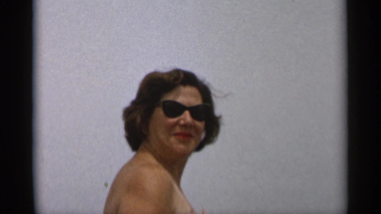 Woman in dark sunglasses and strapless swimsuit posing outdoors against a pale backdrop.