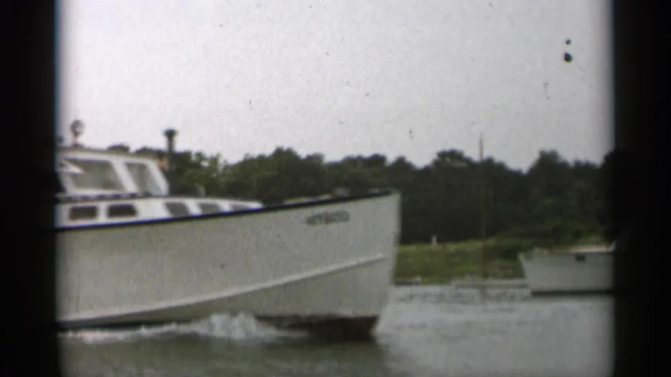 Cape Cod home-movie frame of a white cabin cruiser under way, with a wooded shoreline and another small boat in the background.