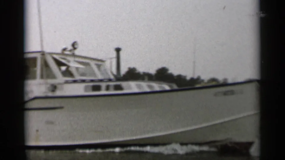 1956 Cape Cod home-movie frame showing a white cabin motorboat cruising on calm water with trees in the background.