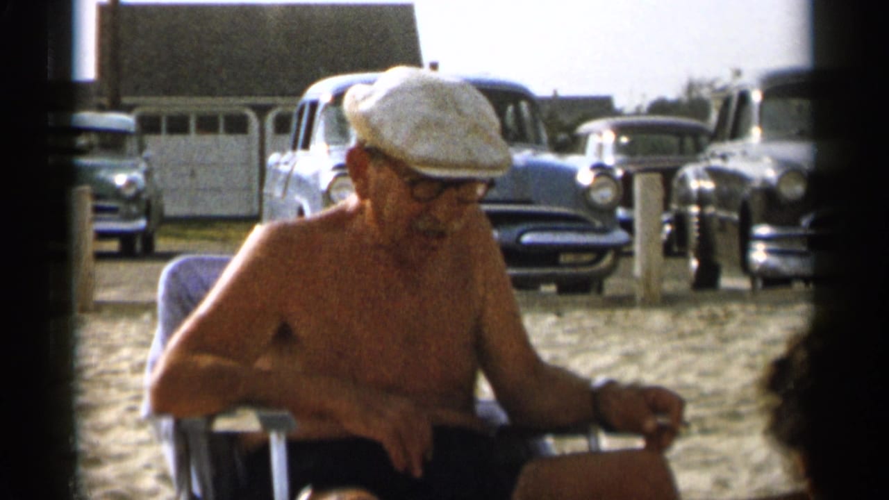 Older shirtless man in a white cap sits on a beach chair with vintage cars behind him.