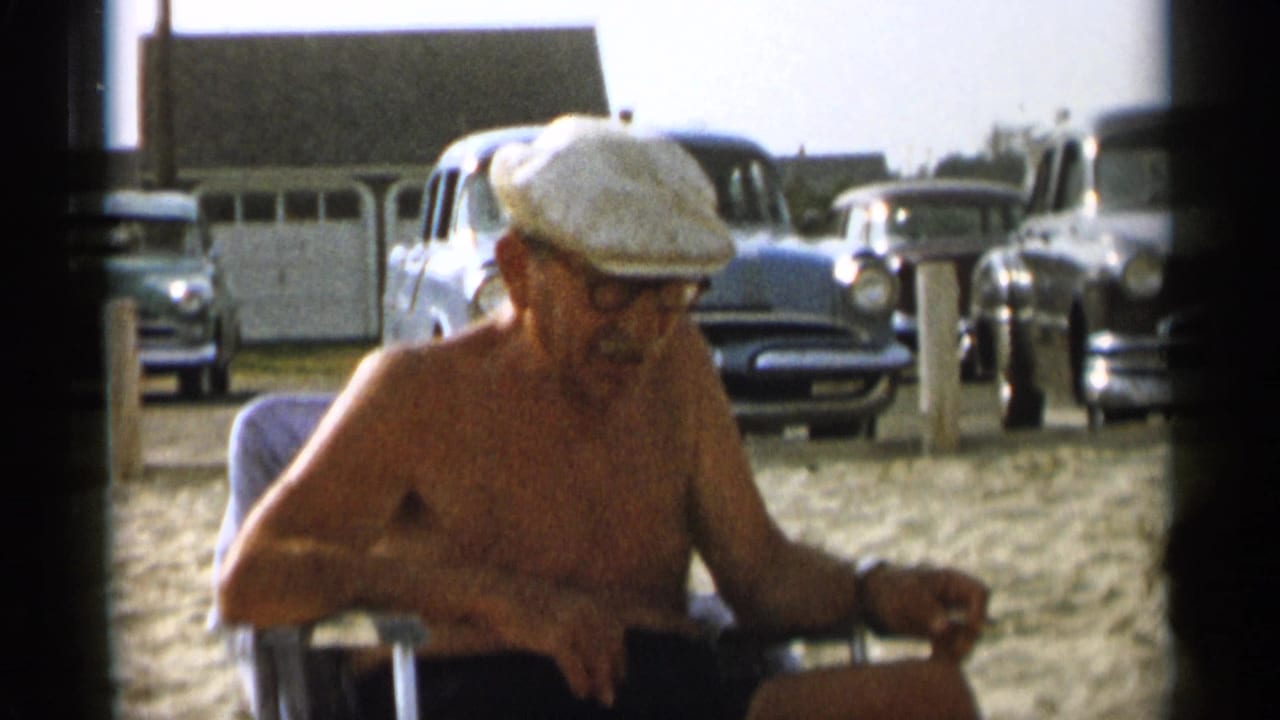 Older man in a white cap looks downward while seated near cars on the beach.