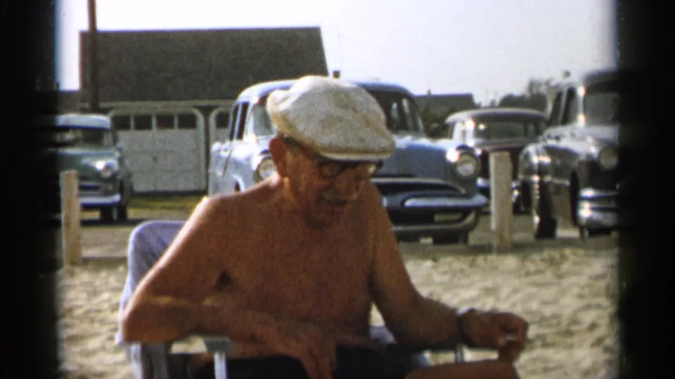 A candid 1956 Cape Cod frame shows the older man seated on the beach, smiling slightly toward someone off-camera.