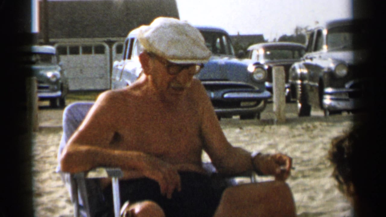 In 1956 Cape Cod footage, an elderly shirtless man wearing glasses and a white flat cap rests in a chair on a sandy beach near parked cars.