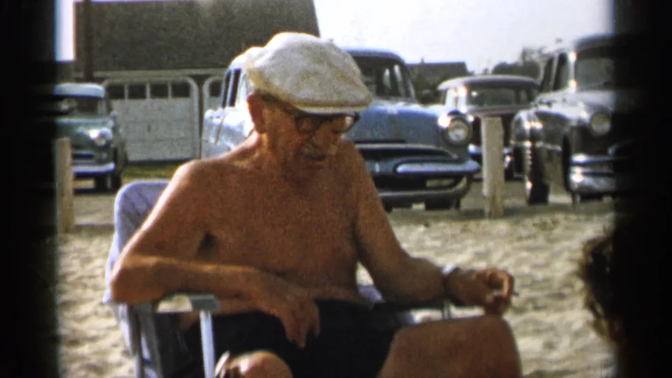The elderly man remains seated in a folding chair on the Cape Cod beach, with classic mid-1950s cars visible in the background.