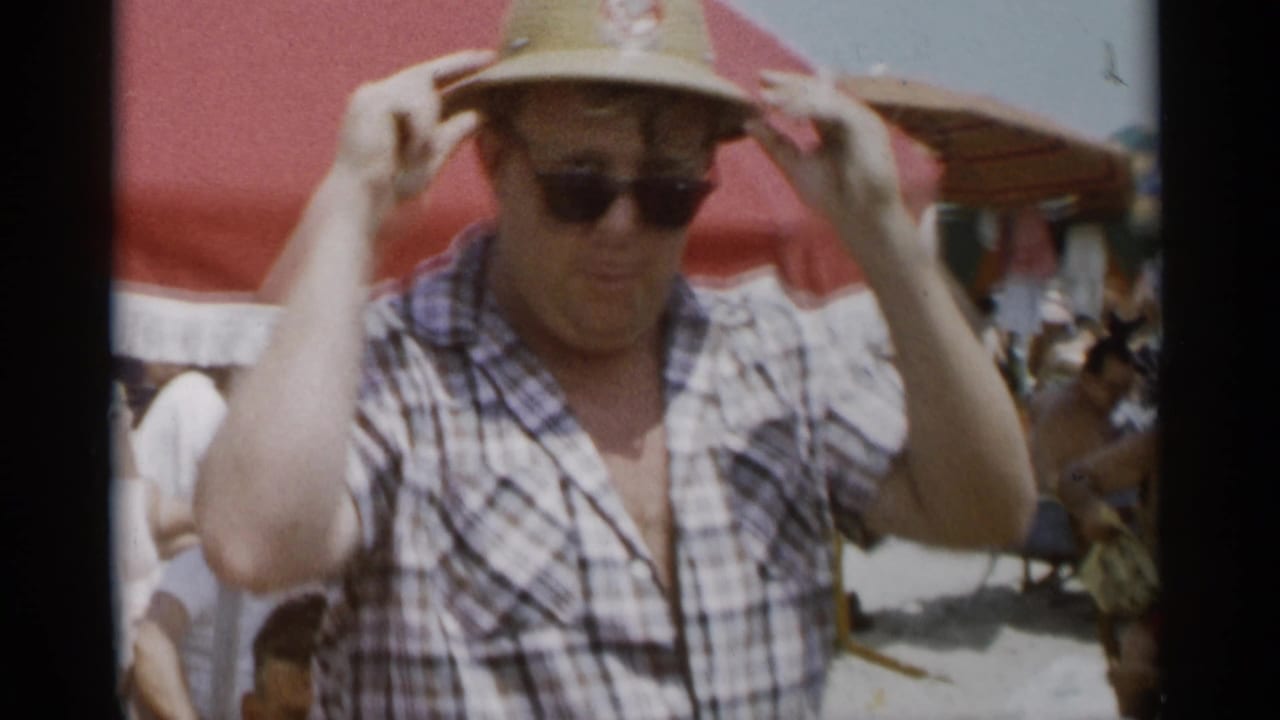 Man in sunglasses adjusts a straw hat at a crowded beach.