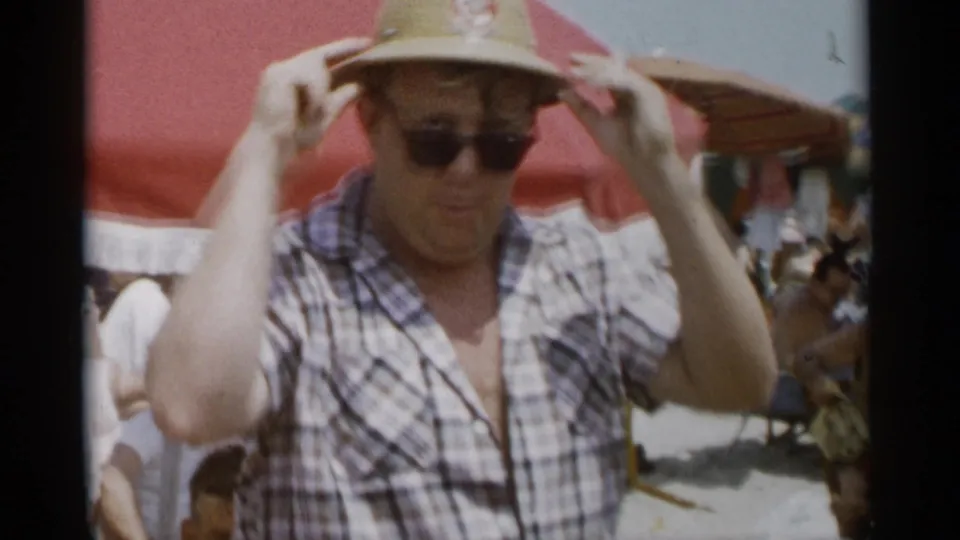 At Jones Beach in 1956, a man in a plaid shirt and sunglasses adjusts his straw hat near red beach umbrellas.