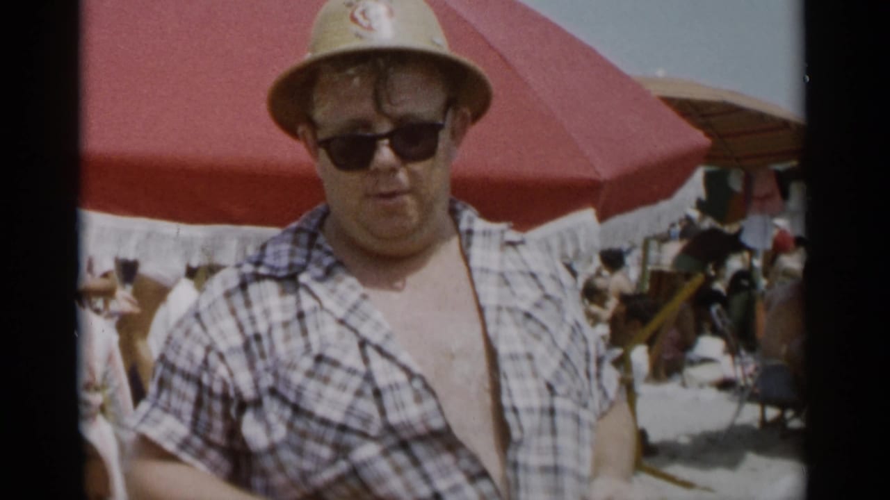 The man stands facing the camera at Jones Beach, wearing sunglasses, a straw hat, and an open plaid shirt.