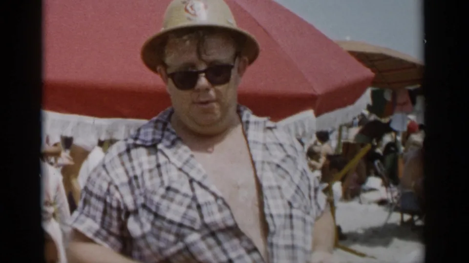 The man stands facing the camera at Jones Beach, wearing sunglasses, a straw hat, and an open plaid shirt.