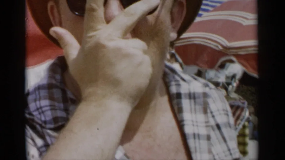 A close handheld frame shows the man partly obscuring his face with his hand under red beach umbrellas.