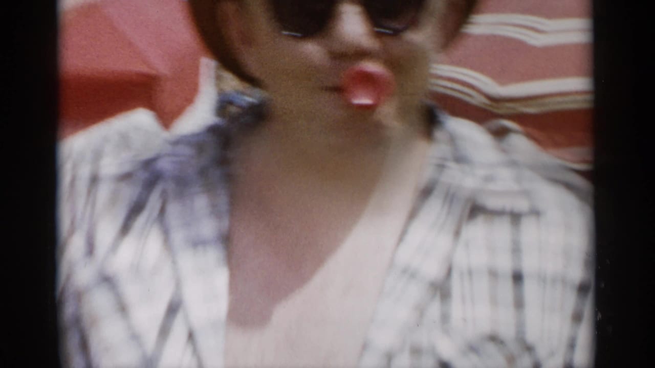 Close view of a woman in sunglasses with a red object near her mouth under a striped beach umbrella.