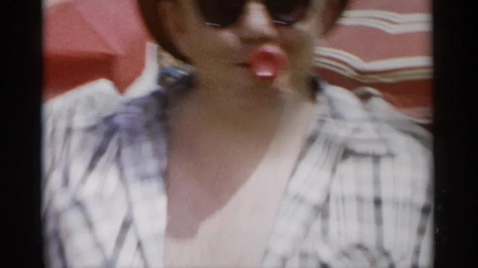 At Jones Beach in 1956, a woman in sunglasses faces the camera with a red object near her mouth beneath a striped beach umbrella.
