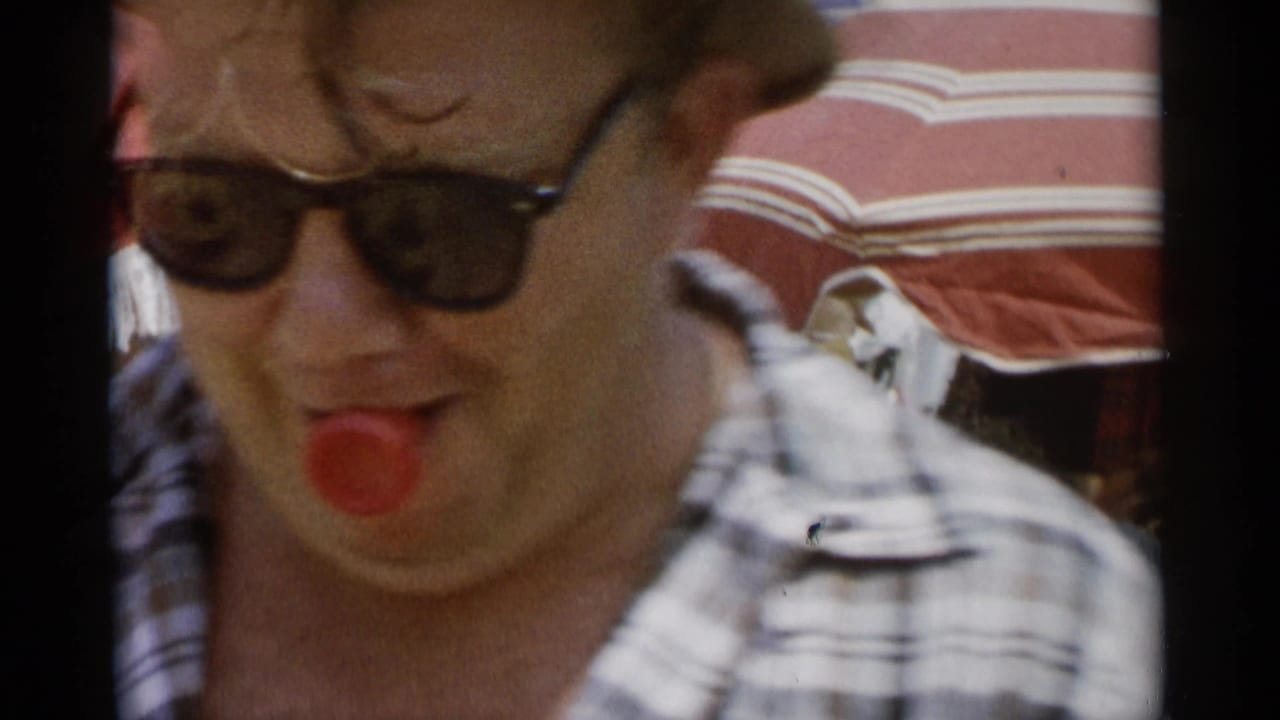 At Jones Beach in 1956, a man in dark sunglasses makes a goofy face with a red object or tongue visible beneath a striped beach umbrella.