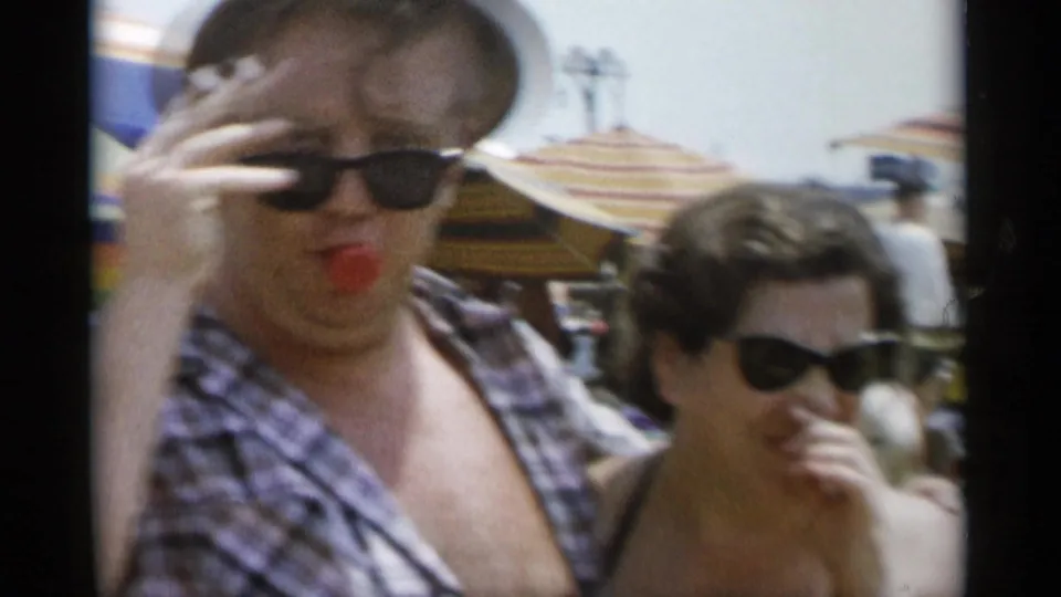 At Jones Beach in 1956, a man in sunglasses touches his face as a woman in sunglasses stands beside him among striped umbrellas and beachgoers.