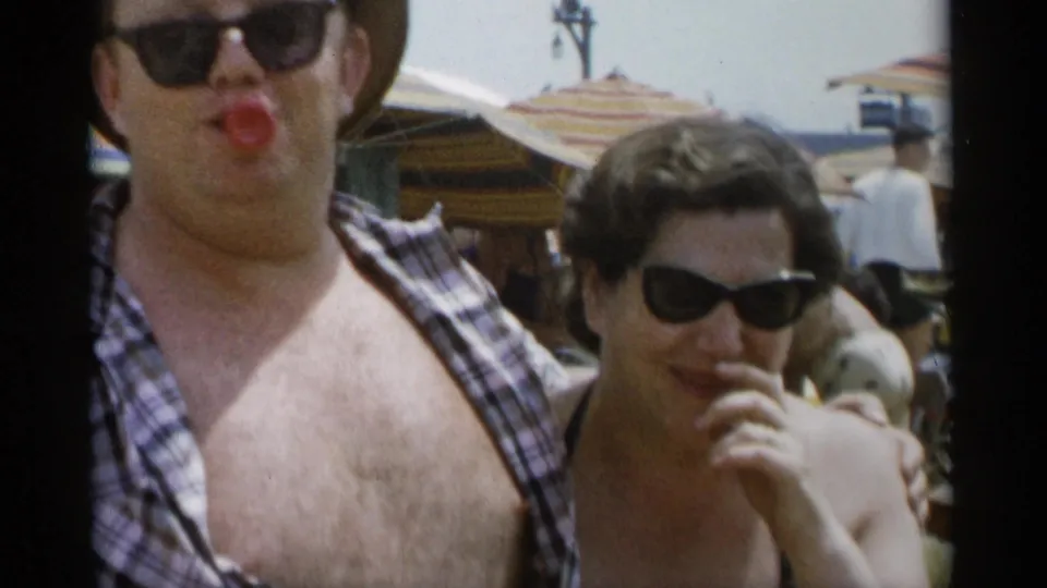 At Jones Beach in 1956, a man with an open plaid shirt and sunglasses poses beside a woman in sunglasses with beach umbrellas behind them.