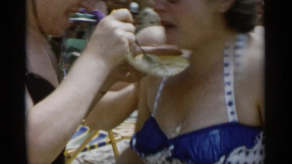 Close home-movie frame of a woman eating from a plate at Jones Beach, with beach chairs and sunbathers behind her.