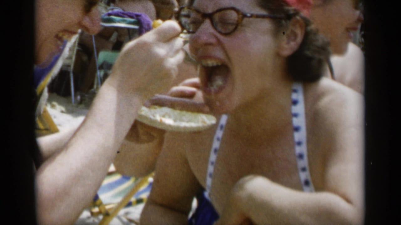 Laughing woman in glasses being fed cake on a crowded beach.