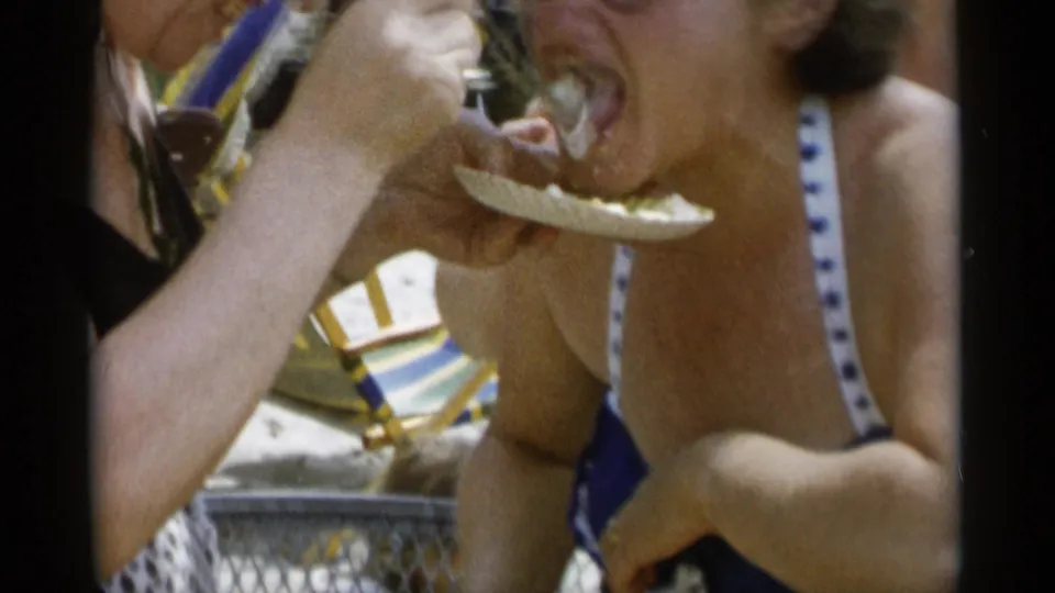 In a close handheld frame, the woman opens her mouth as a friend feeds her cake during a beach outing.