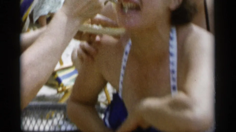 The woman continues laughing as the plate is held near her face, with beach seating and visitors behind her.