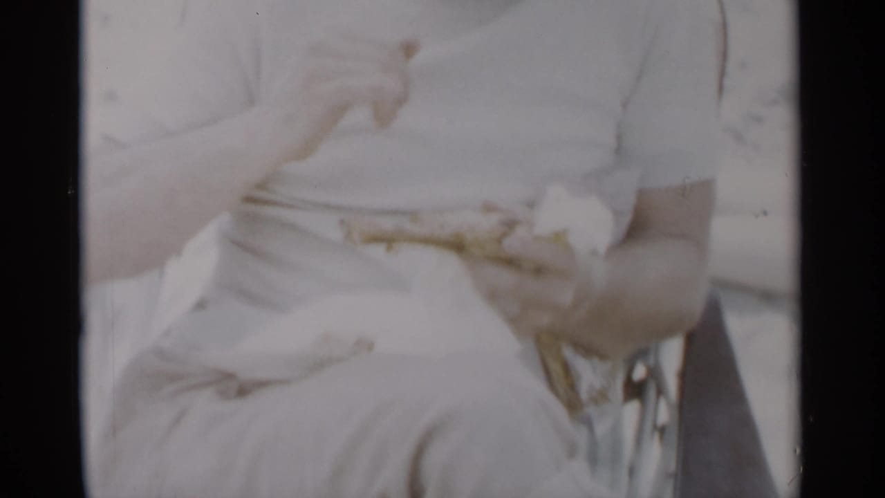 At Jones Beach in 1956, a seated man in a white shirt holds food in a bright, overexposed beach frame.