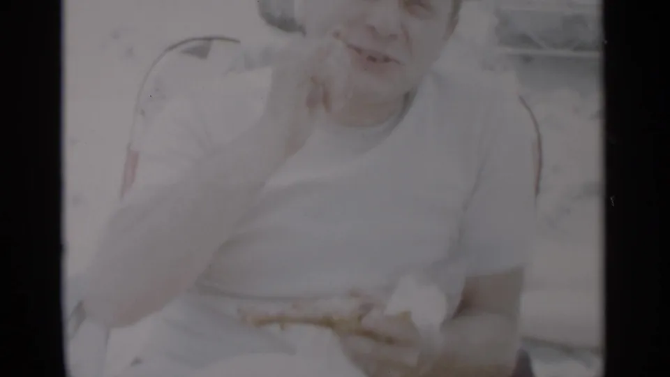 At Jones Beach in 1956, a man in a white shirt appears to eat while seated in a washed-out home-movie beach frame.