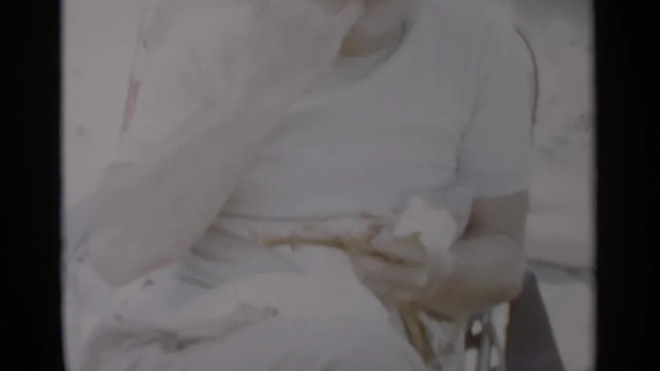 At Jones Beach in 1956, a seated man in a white shirt holds food near a beach chair in a softly exposed 8mm frame.