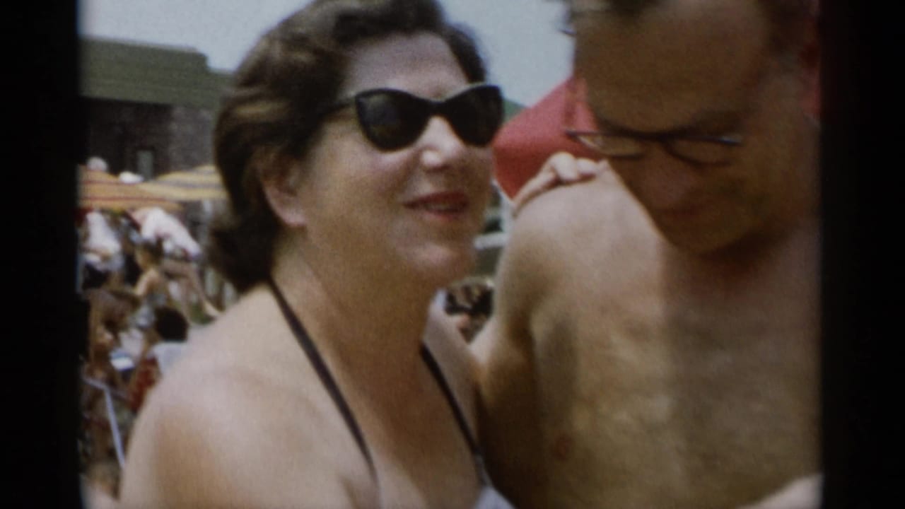 Woman in sunglasses smiles beside a shirtless man at a crowded beach.