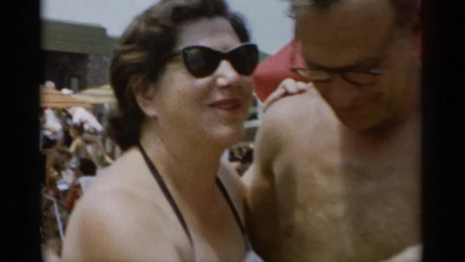 At Jones Beach in 1956, a woman in dark sunglasses smiles with her arm around a shirtless man amid beachgoers.