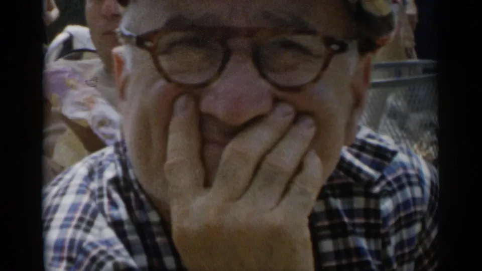 At Jones Beach in 1956, a man wearing glasses and a plaid shirt laughs toward the camera with his hand over his mouth.
