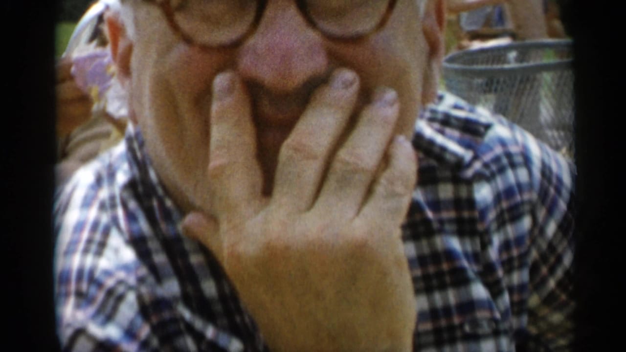Older man in round glasses covers his mouth while smiling close to the camera.