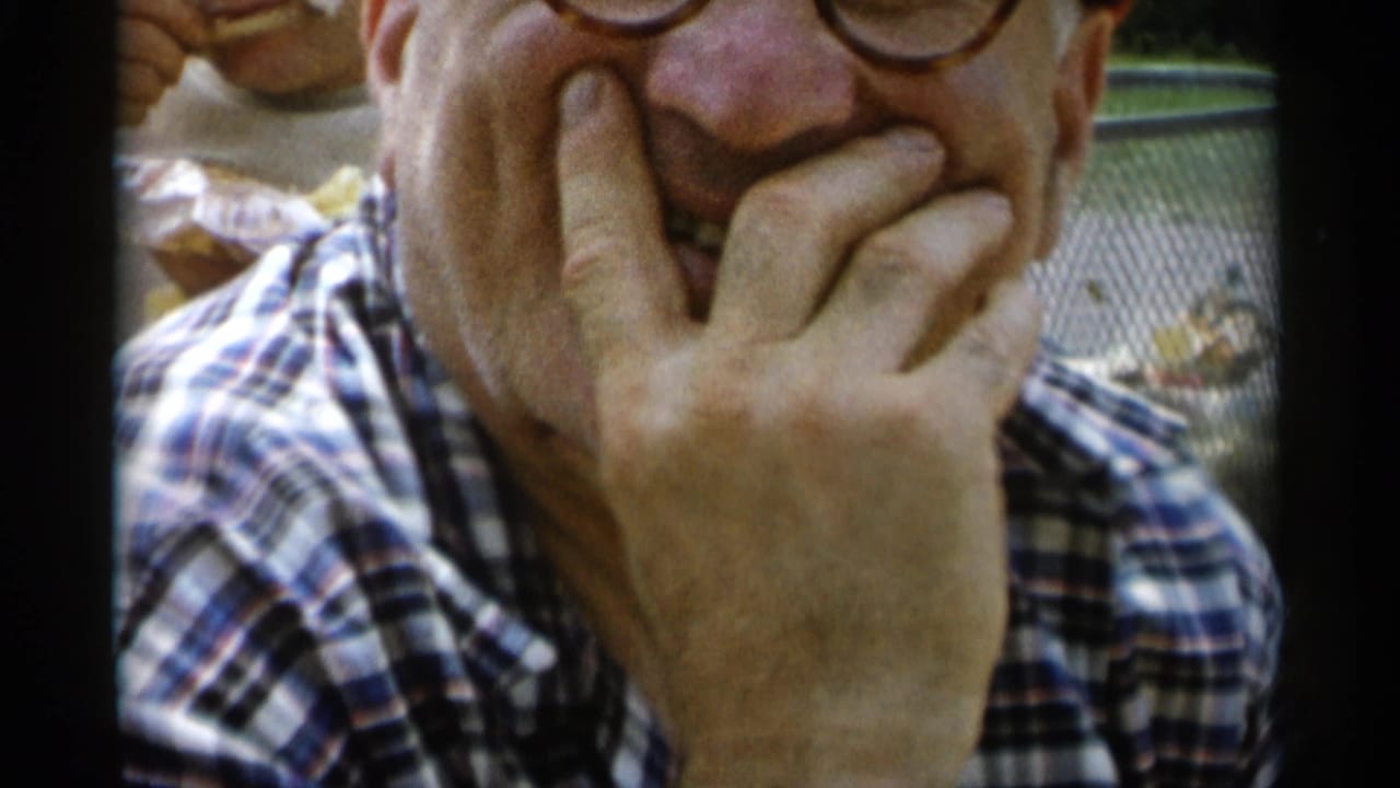 Older man in glasses smiles with his hand against his face near a picnic table.