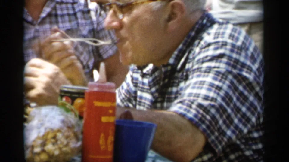 Side view from the 1956 Jones Beach picnic shows the older man eating beside red and blue cups, packaged snacks, and another plaid-shirted adult.