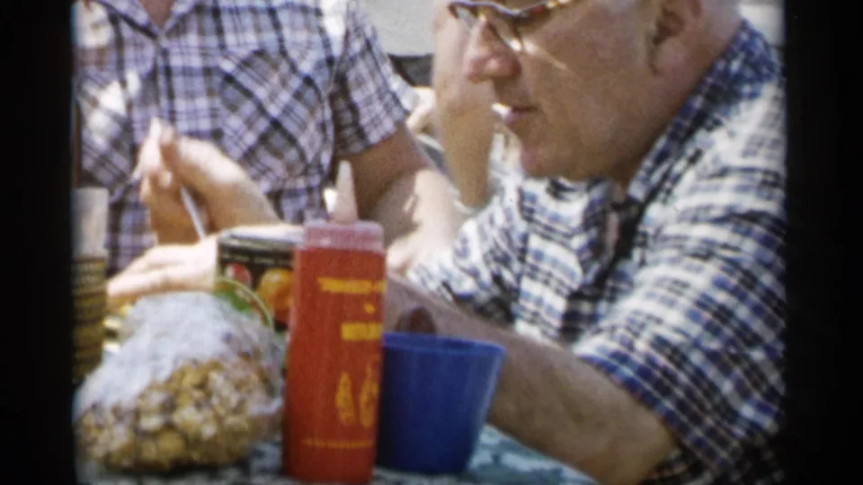 The final 1956 Jones Beach frame shows the older man seated at the picnic table with cups, food packaging, and another adult nearby.