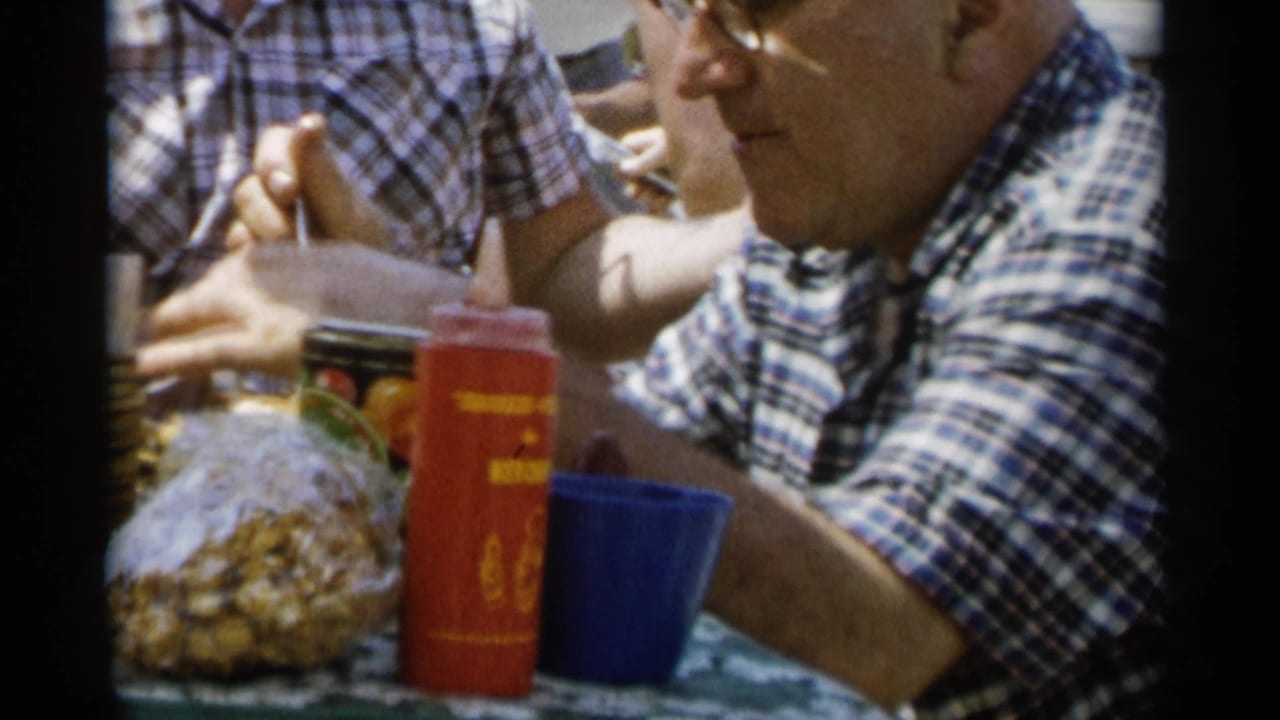Man in glasses seated at a picnic table with cups, food containers, and another plaid-shirted person nearby.