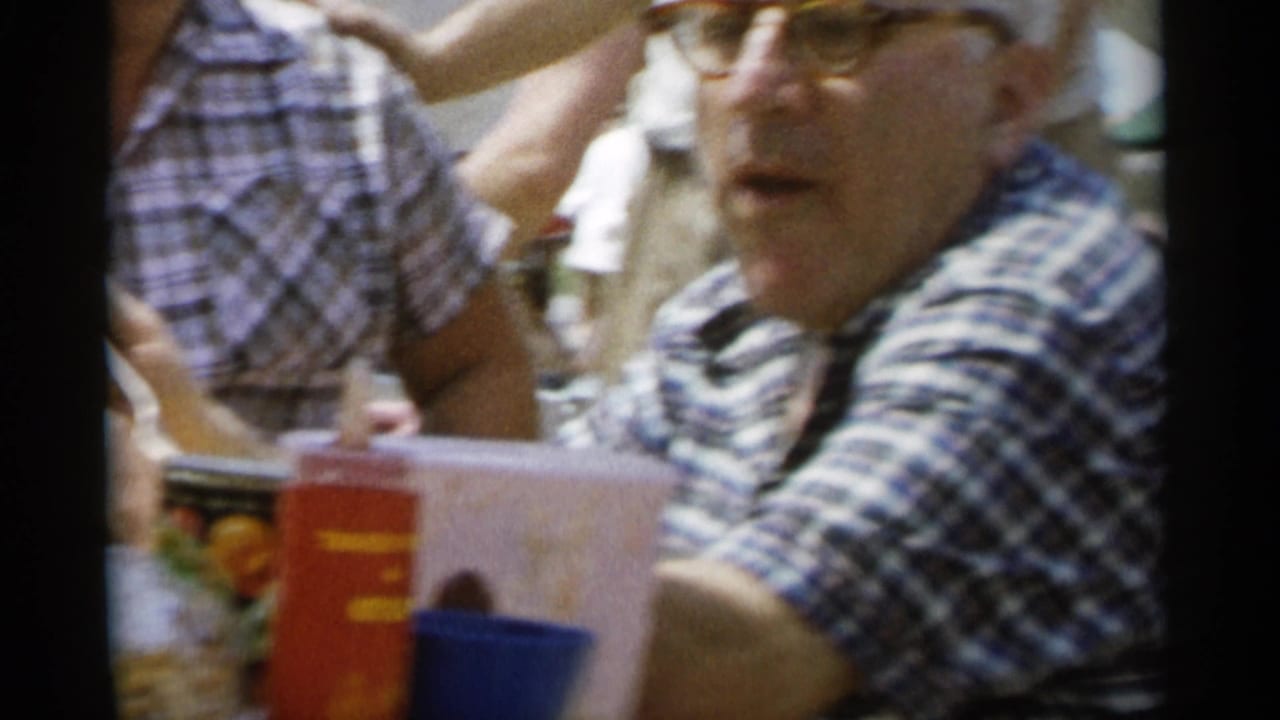 Man in glasses at an outdoor picnic table behind a red bottle, blue cup, and pale container.