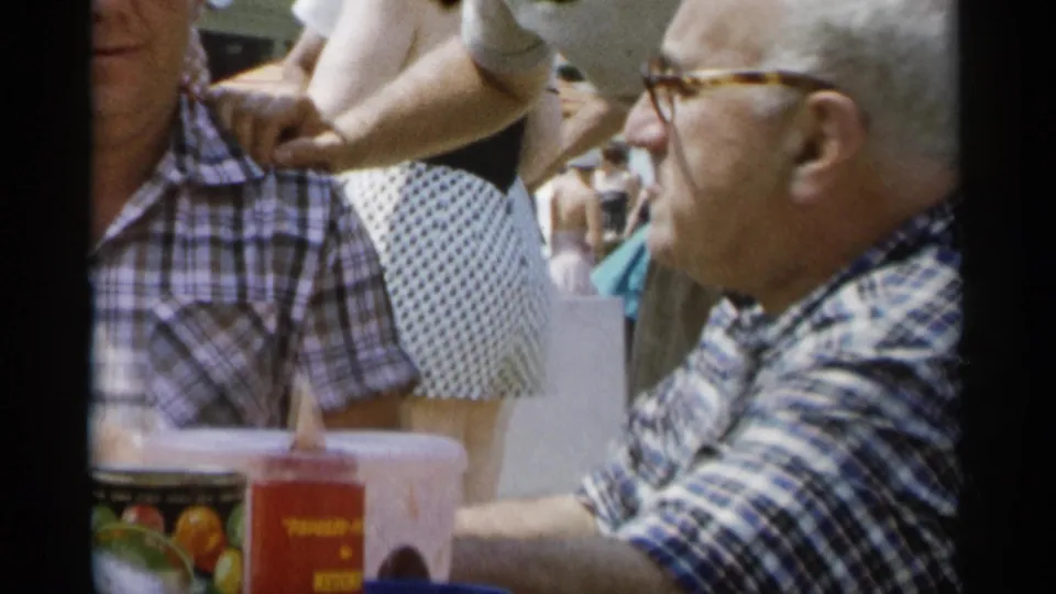 At Jones Beach in 1956, two adult men in plaid shirts sit close together at a sunny picnic table with food and drink items visible.