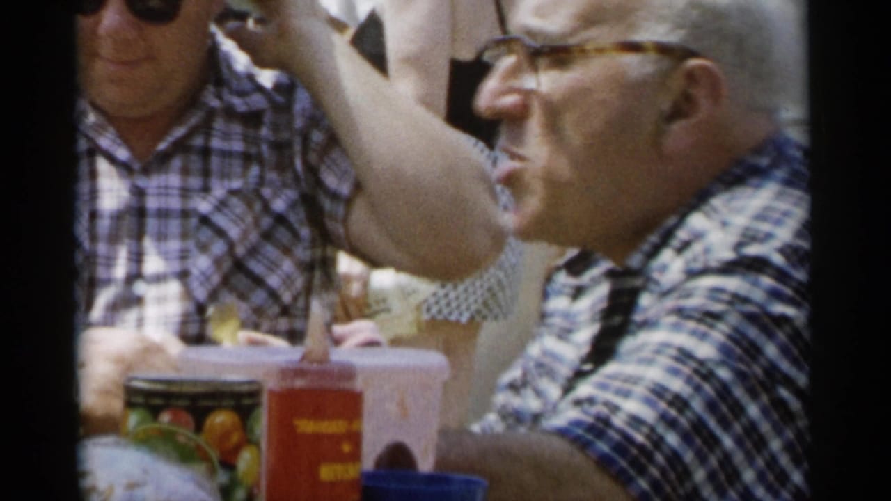 A man in glasses sits beside another man at a picnic table with cups and a red bottle nearby.