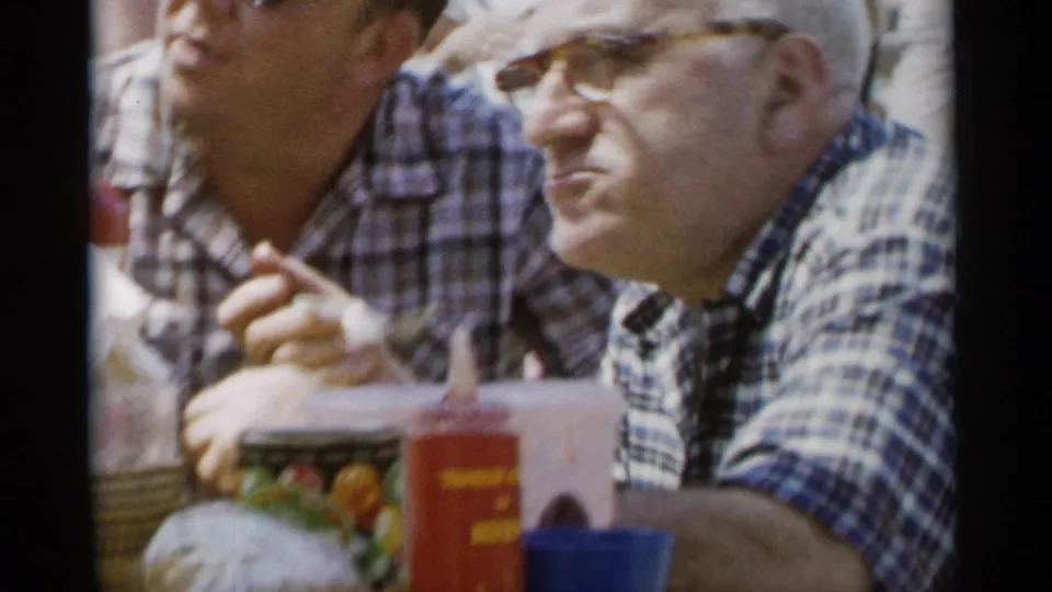 At the Jones Beach picnic, a man in glasses and a plaid shirt faces the camera as another adult sits beside him near cups and food containers.