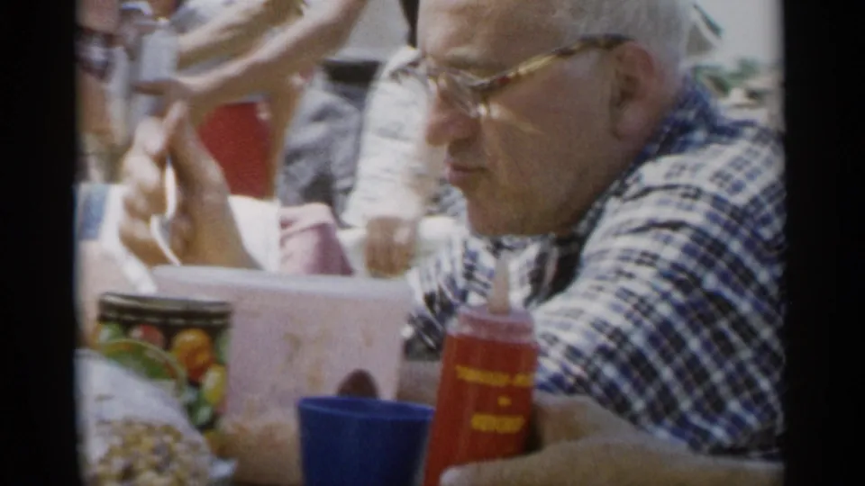 In this 1956 Jones Beach frame, a man in glasses holds a spoon at a picnic table with cups, a red condiment bottle and food items nearby.