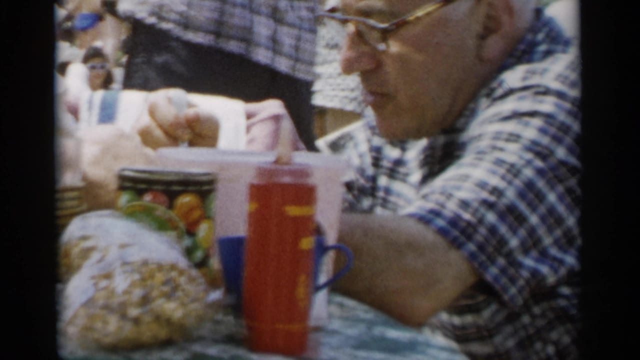 Older man in plaid shirt eating beside a red thermos and table snacks.