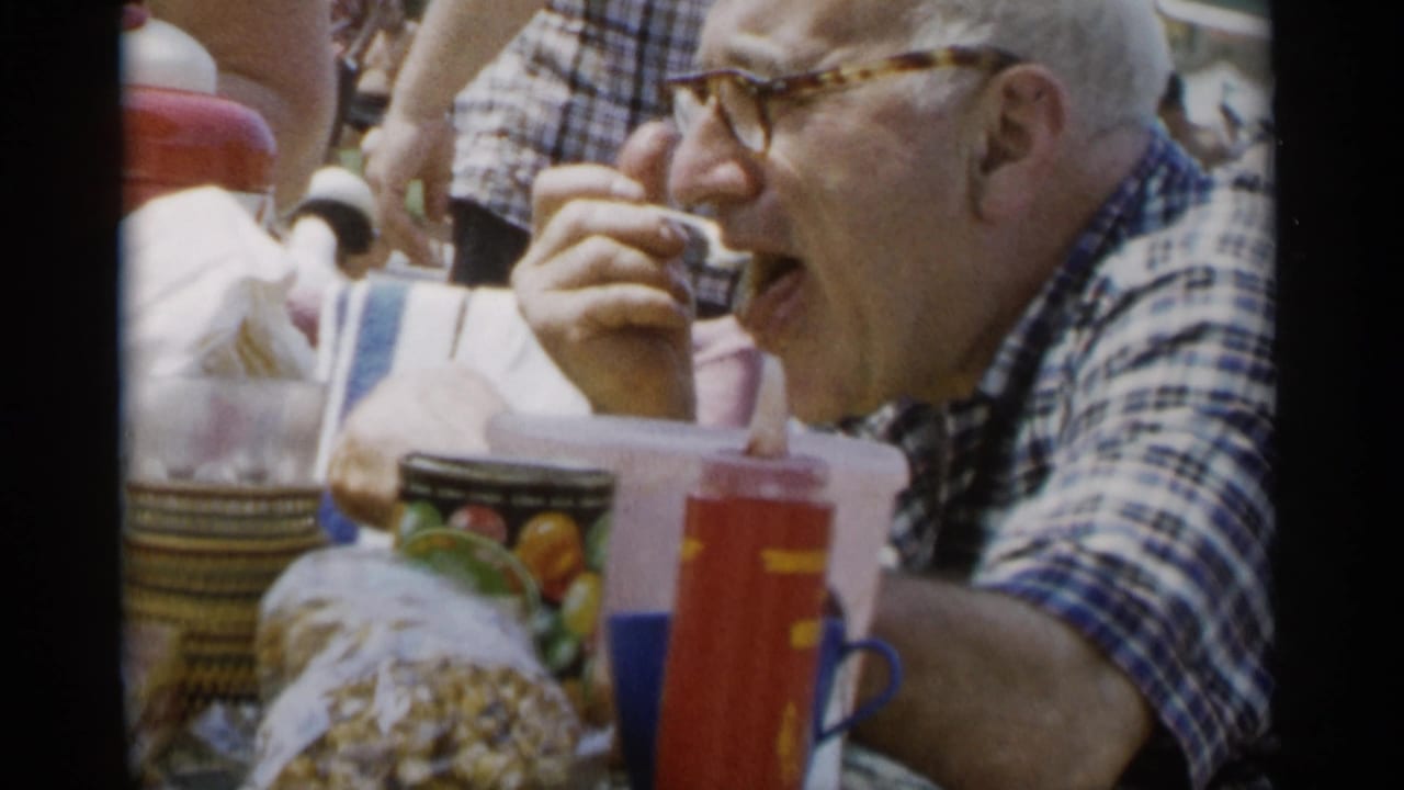 Older man brings food to his mouth at an outdoor table with cups and snacks.