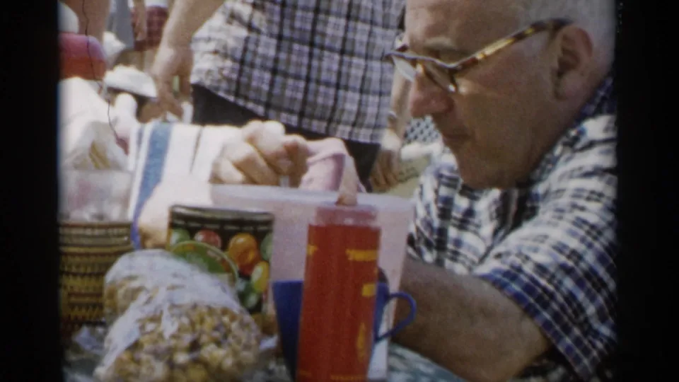 The 1956 Jones Beach home movie frame shows the man eating at a crowded outdoor table with snacks and cups.