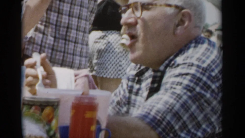 Older man in a plaid shirt chews at a Jones Beach outdoor gathering in 1956, with tableware and people behind him.
