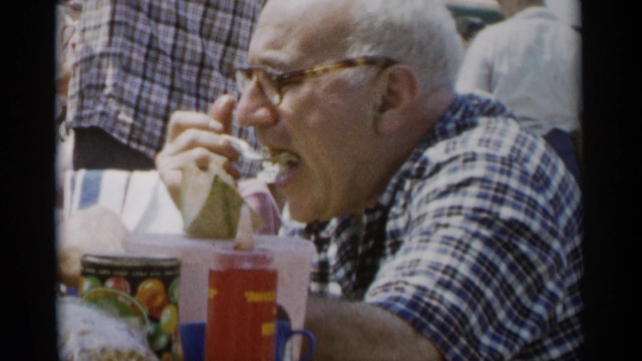 Older man in glasses eating at an outdoor picnic table.