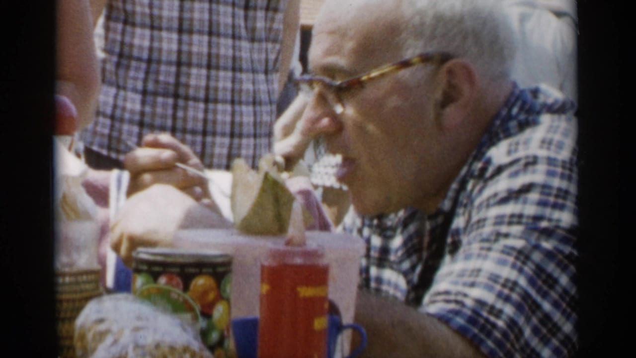 Older man in profile eating beside picnic table items.