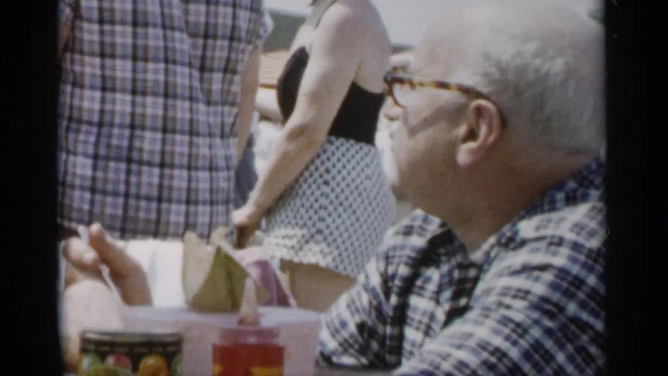 The seated older man looks up from the picnic table as standing guests in summer clothing pass behind him at Jones Beach.